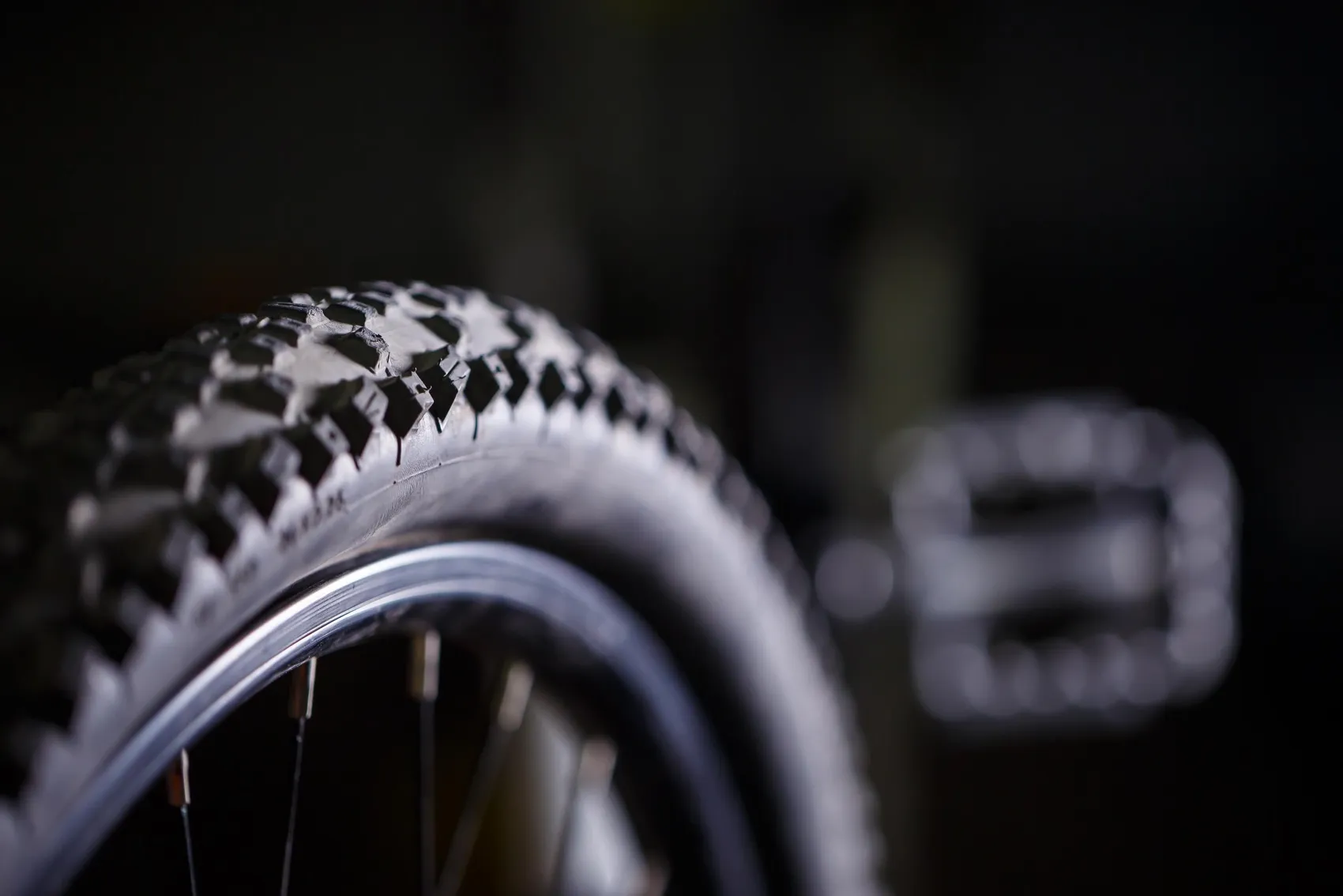 Nahaufnahme eines Mountainbike-Reifens mit grobem Profil und einem Pedal vor dunklem Hintergrund.