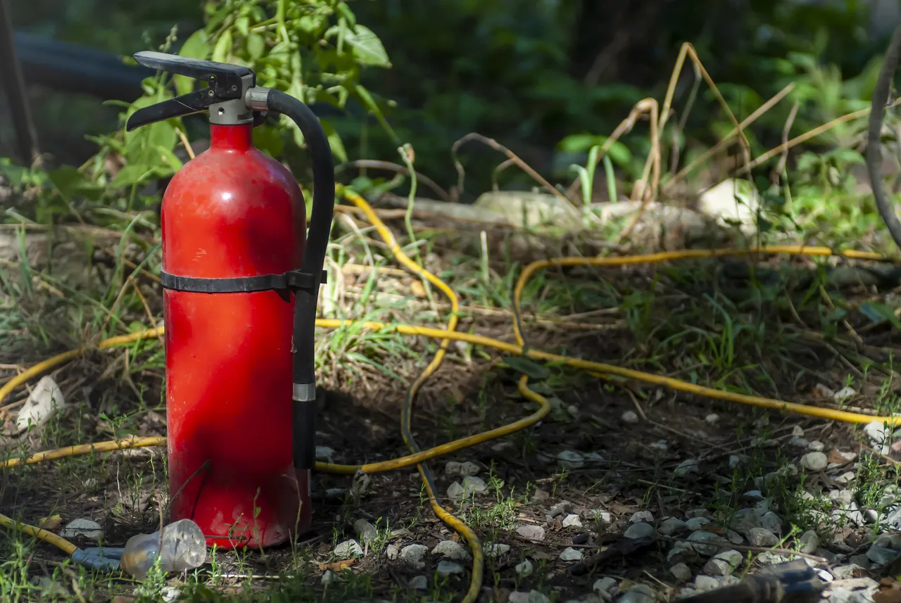 Roter Feuerlöscher steht im Freien neben einem gelben Schlauch.