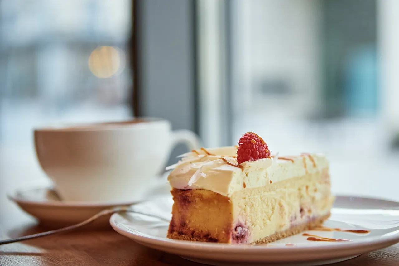 Ein Stück Käsekuchen mit Sahnehaube und einer Himbeere auf einem Teller neben einer Tasse Kaffee.