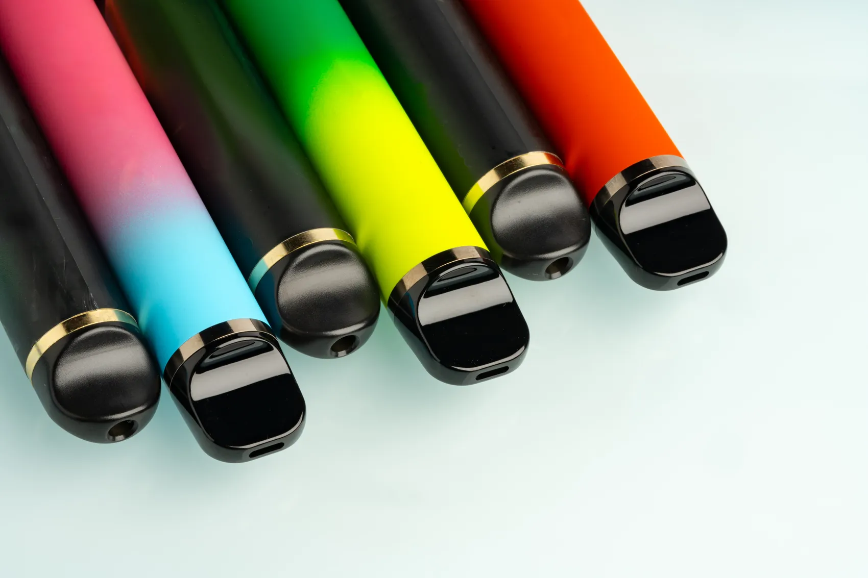 Mehrere bunte E-Zigaretten in verschiedenen Farben und Verläufen liegen diagonal auf einer hellblauen Oberfläche.