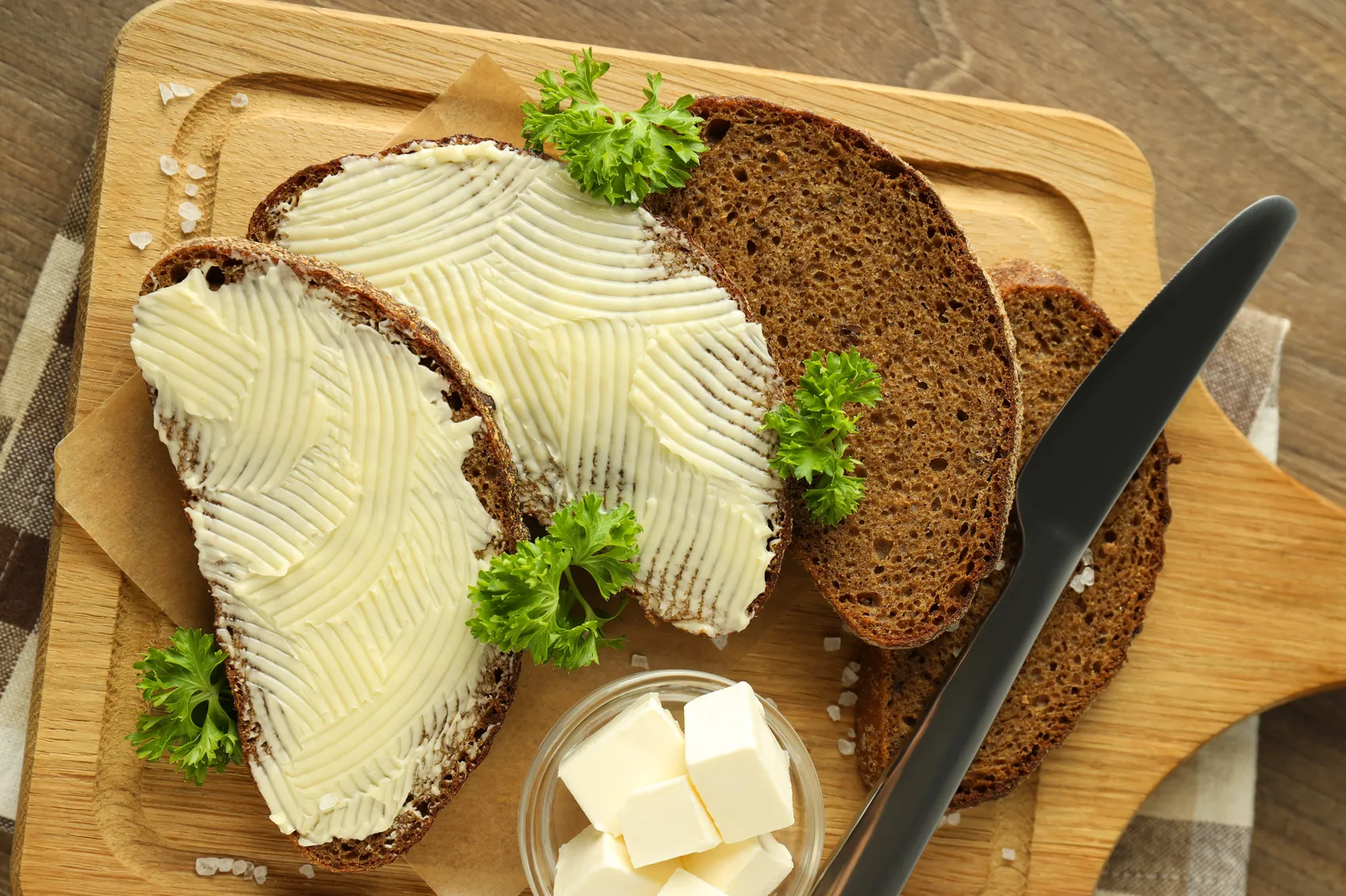 Zwei Scheiben dunkles Brot mit Butter bestrichen und mit Petersilie garniert liegen auf einem Holzbrett neben einem Glas Butterwürfeln und einem Buttermesser.