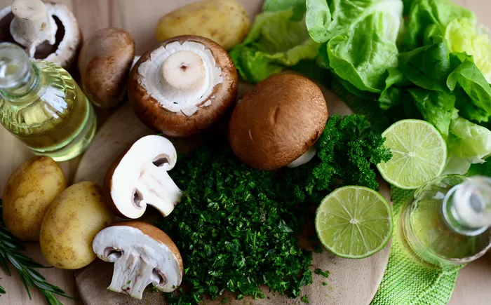 Frische Zutaten wie braune Champignons, Kartoffeln, grüner Salat, Limettenscheiben, gehackte Kräuter und Ölflaschen auf einem Holzbrett.