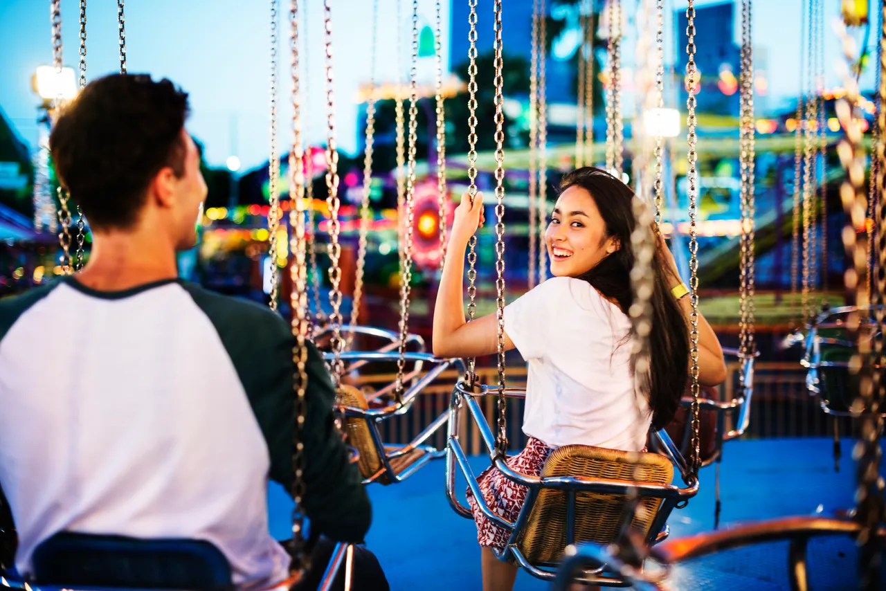 Eine lächelnde junge Frau blickt zu einem jungen Mann in einem Kettenkarussell auf einer hell erleuchteten Kirmes.