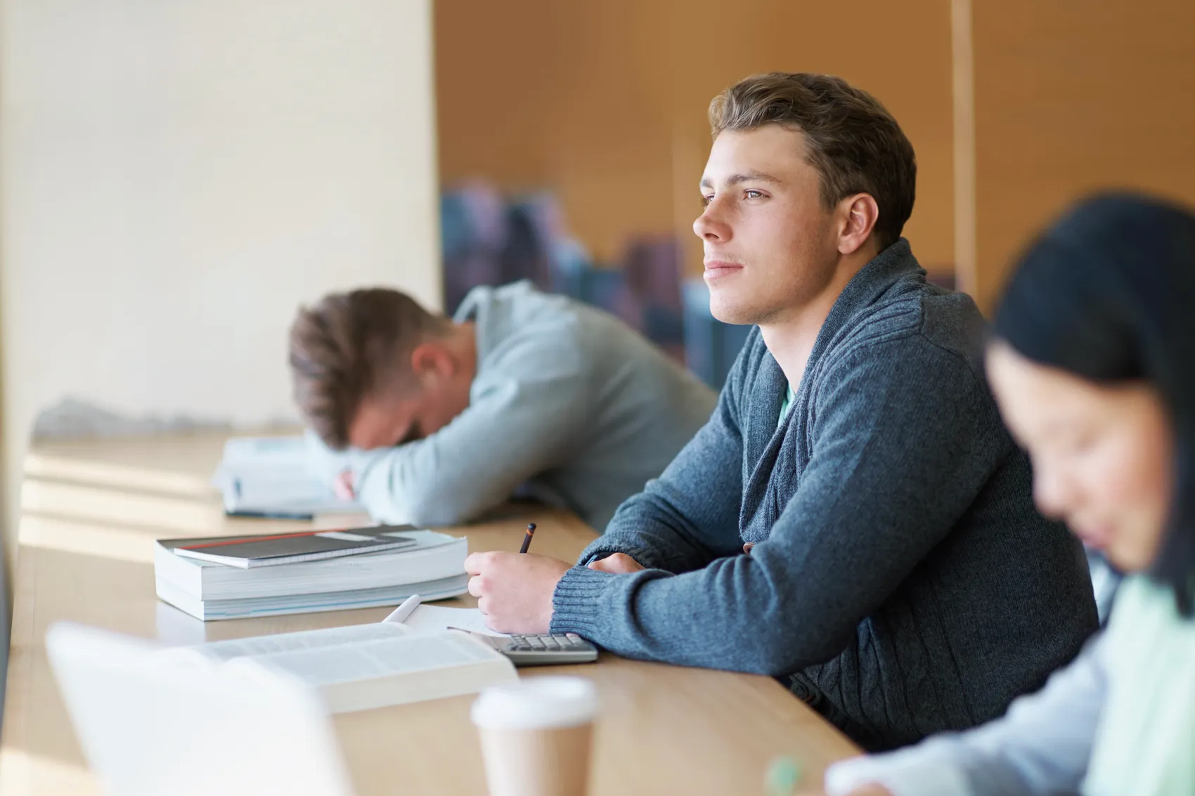 Ein junger Mann blickt während einer Lernsession aufmerksam in die Ferne, während ein Mitschüler erschöpft auf dem Tisch schläft.