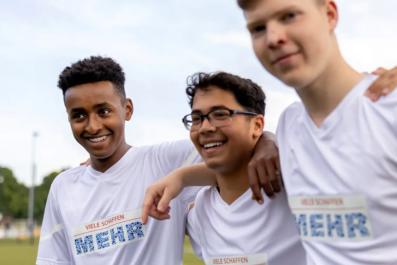Drei lächelnde junge Männer in weißen T-Shirts mit der Aufschrift „VIELE SCHAFFEN MEHR“ stehen mit den Armen umeinander.