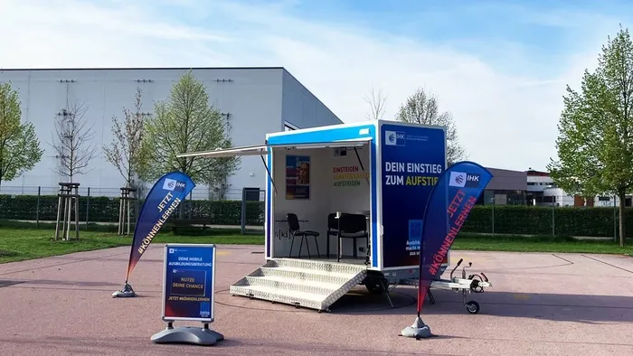 Ein weißer mobiler Beratungsanhänger der IHK mit geöffneter Seite und zwei blauen Bannern mit dem Hashtag #KÖNNENLERNEN steht auf einem gepflasterten Platz.