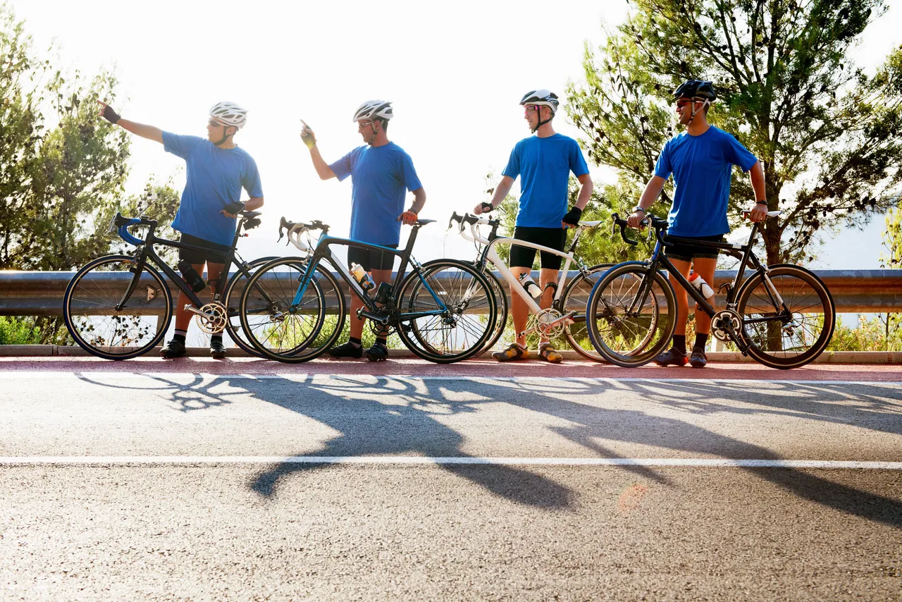 Vier Radfahrer in blauen T-Shirts und Helmen stehen mit ihren Rennrädern am Straßenrand.