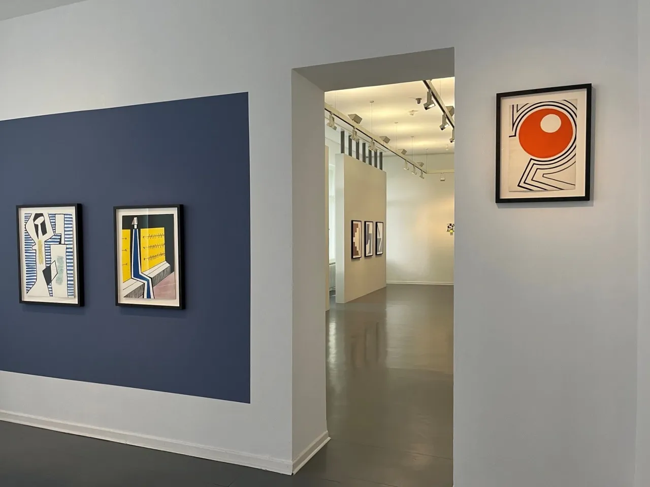 Gerahmte abstrakte Gemälde hängen an weißen und blauen Wänden in einem hellen Galerieraum.