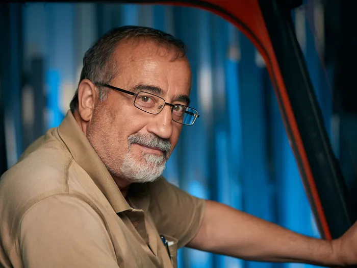 Erdal Ilgan, ein Mann mit graumeliertem Haar und Brille, lächelt in die Kamera. Im Hintergrund ist ein verschwommener Arbeitsbereich mit blauen und roten Elementen zu sehen.