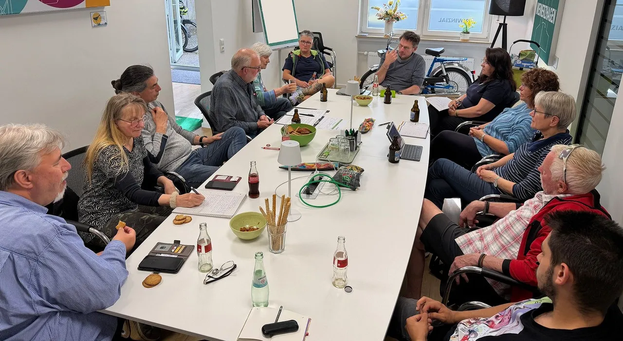 Eine Gruppe von etwa zehn Personen sitzt um einen großen Tisch in einem hellen Raum und diskutiert. Auf dem Tisch stehen Getränke, Snacks und Smartphones. Im Hintergrund sind Fahrräder und ein Ergometer zu sehen.