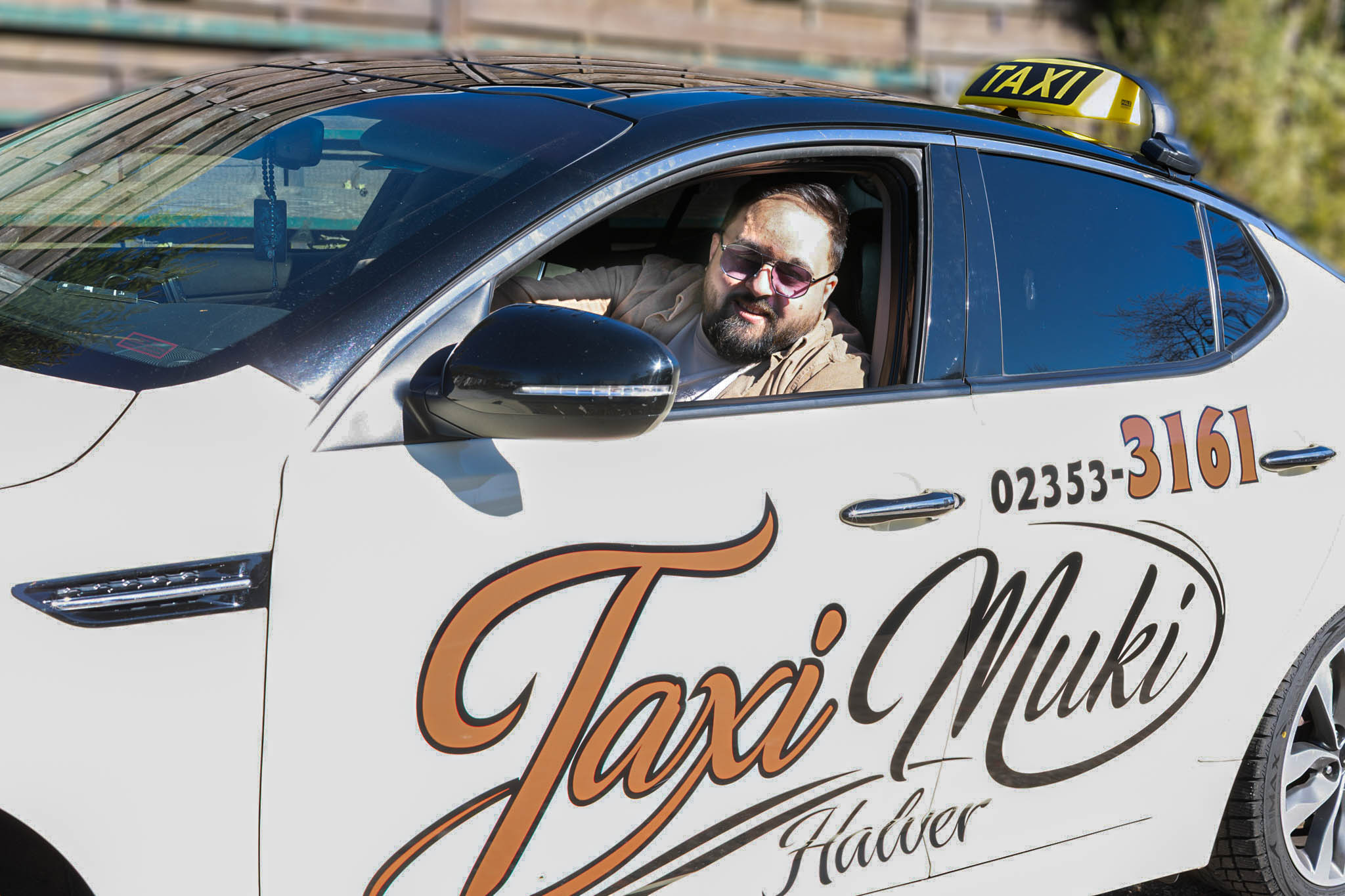Ein Taxifahrer lächelt aus dem Fenster seines weißen Fahrzeugs. Auf der Tür steht in geschwungenen Lettern „Taxi Muki Halver“ und eine Telefonnummer.