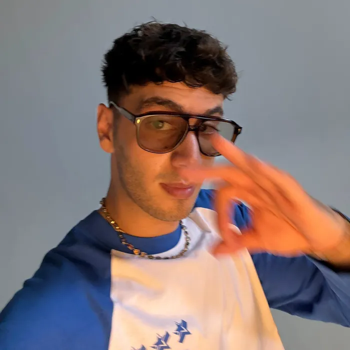 Junger Mann mit dunklen Locken und Brille macht ein OK-Handzeichen vor einem grauen Hintergrund.