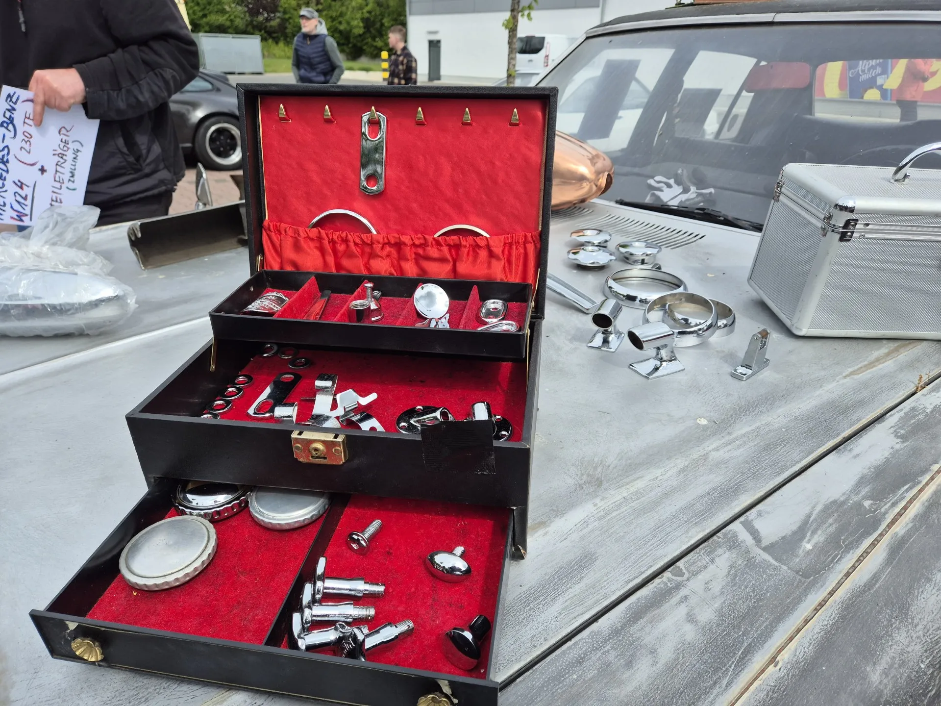 Geöffneter Koffer mit verschiedenen Chromteilen und Werkzeug auf einer Motorhaube eines Oldtimers. Im Hintergrund weitere Oldtimer und Personen.