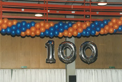 Luftballons in Orange und Blau sowie silberne Folienballons in Form der Zahl 100 hängen vor einer braunen Wand.