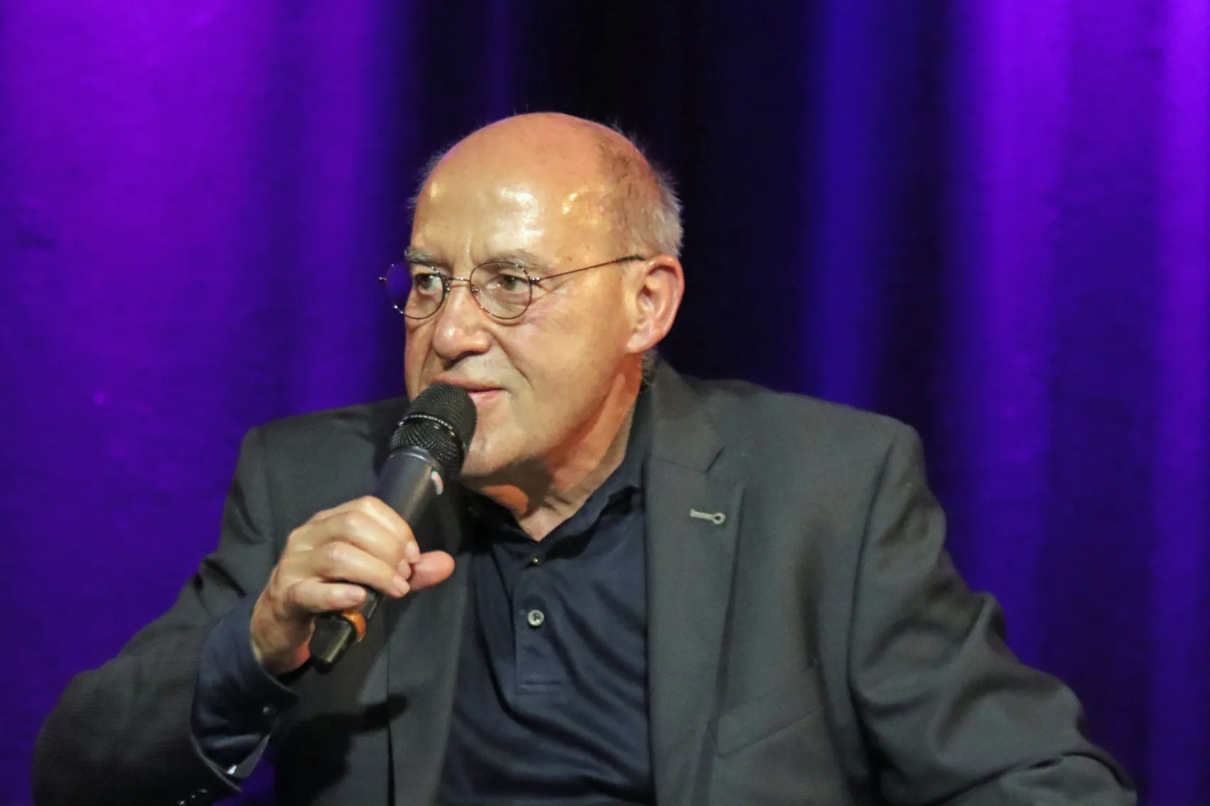 Gregor Gysi spricht in ein Mikrofon vor einem violetten Vorhang.