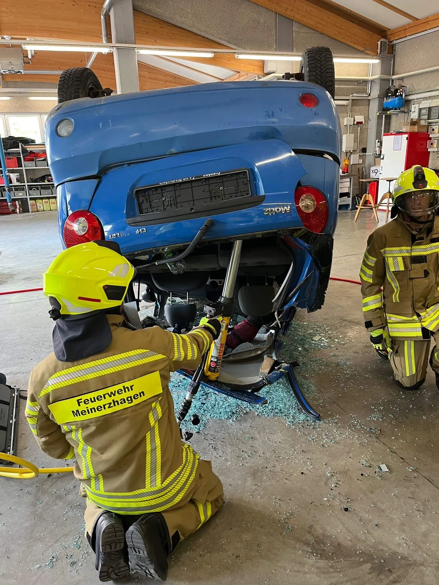 Feuerwehrleute bei der Bergung eines auf dem Dach liegenden blauen Kleinwagens. Sie verwenden hydraulische Rettungsgeräte, während Glassplitter auf dem Boden liegen.