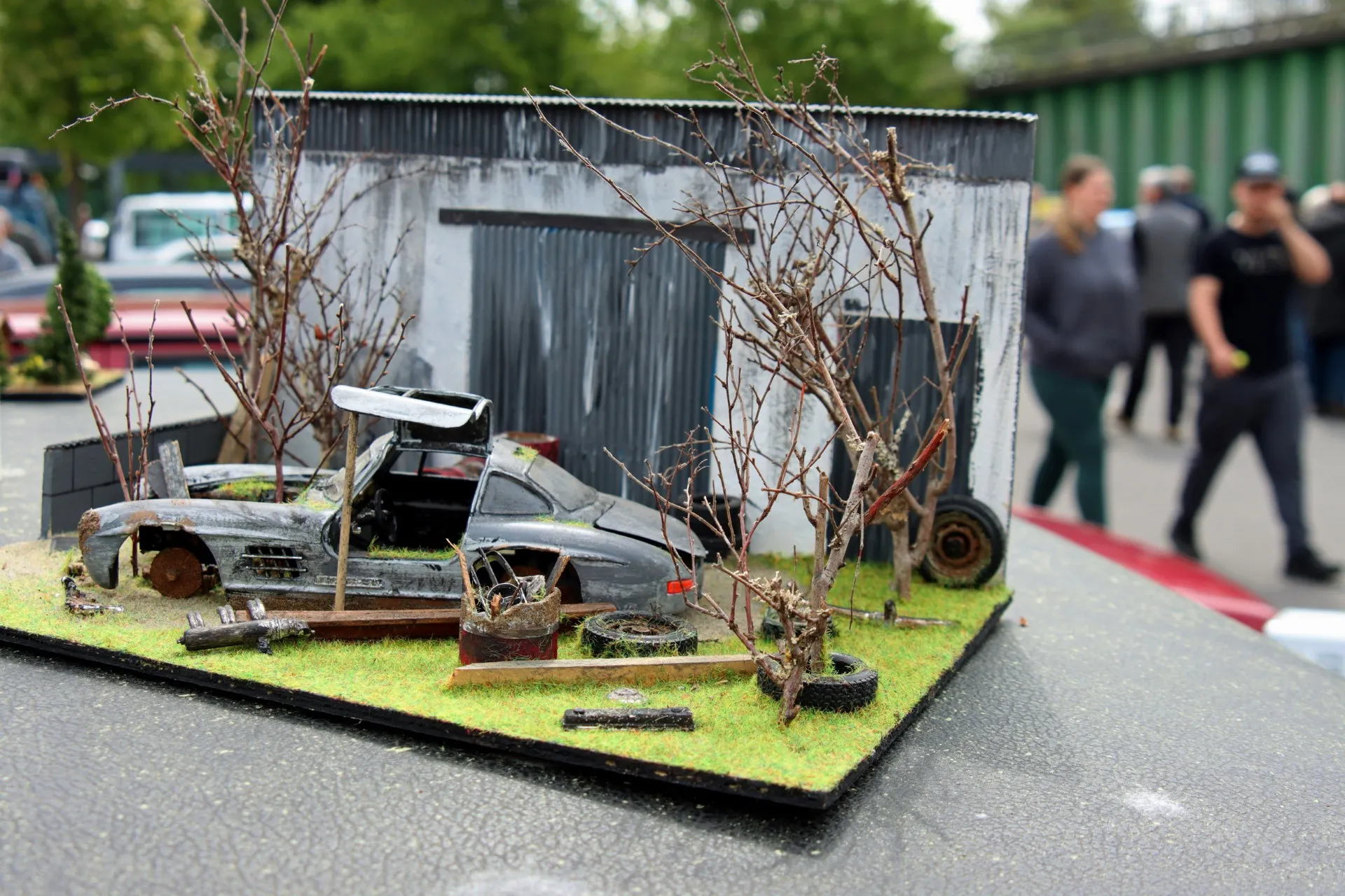 Detailliertes Modell eines verlassenen Autos vor einer verfallenen Lagerhalle, umgeben von Bäumen und Gras.