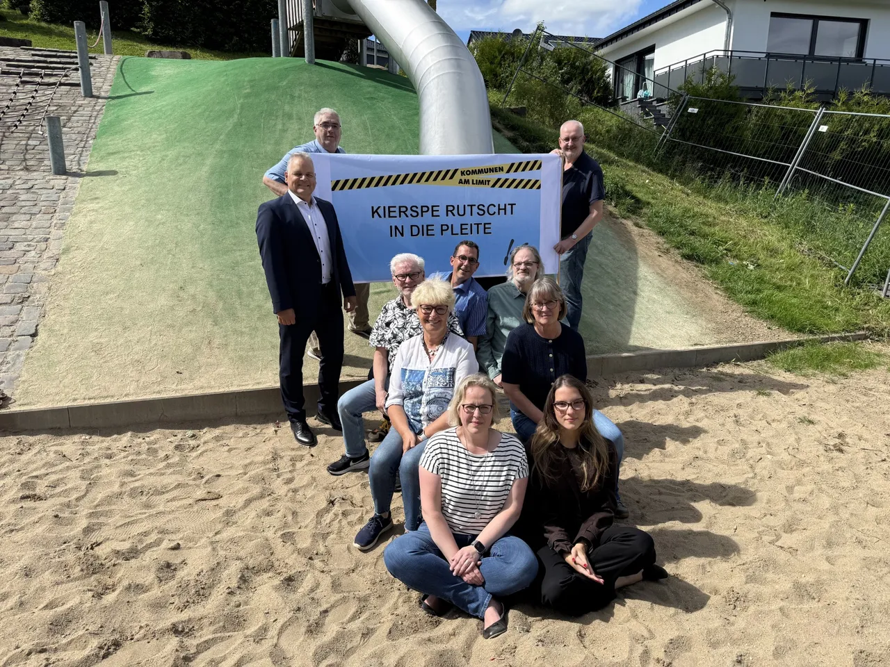 Eine Gruppe von Lokalpolitikern posiert vor einer Sandfläche mit einer Rutsche. Sie halten ein Banner mit der Aufschrift „Kierspe rutscht in die Pleite“ und demonstrieren so gegen die steigende Finanzlast der Gemeinde.