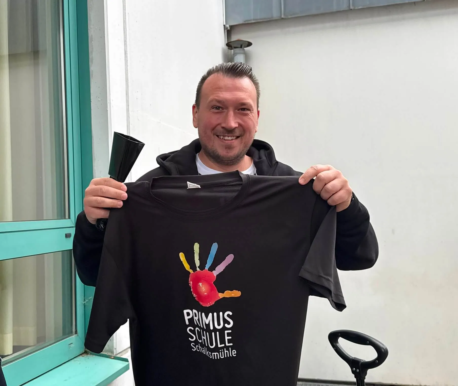 Bürgermeister Christian Breddermann hält ein schwarzes T-Shirt mit einem bunten Handabdruck und dem Aufdruck „PRIMUS SCHULE Schalksmühle“ in die Kamera.