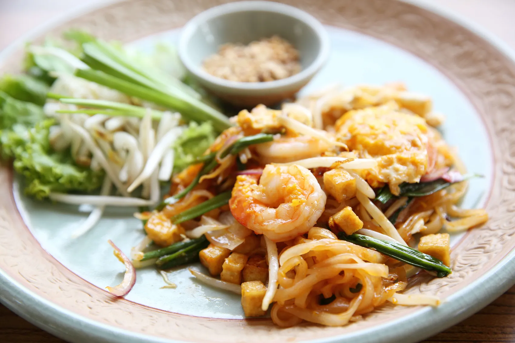 Pad Thai mit Garnelen, Tofu und Sprossen auf einem verzierten Teller, dazu eine kleine Schale mit Gewürzen und frische Kräuter.