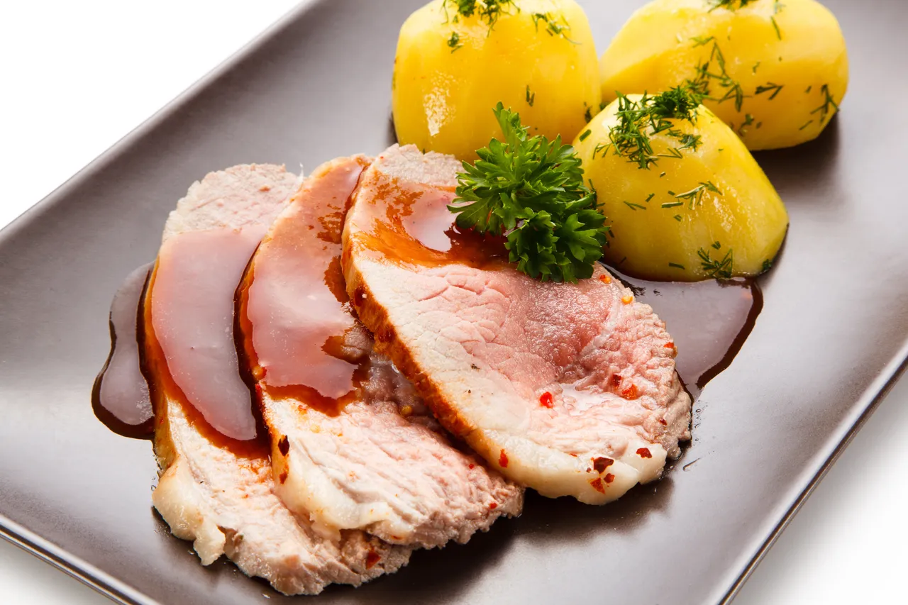 Auf einem dunklen Teller liegen Scheiben Schweinebraten neben drei Salzkartoffeln, garniert mit Petersilie. Eine Soße umrahmt das Fleisch.