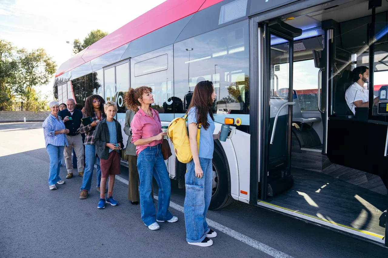 Eine Gruppe von Menschen unterschiedlichen Alters und mit unterschiedlicher Hautfarbe wartet an einer Bushaltestelle auf den Einstieg in einen Stadtbus.