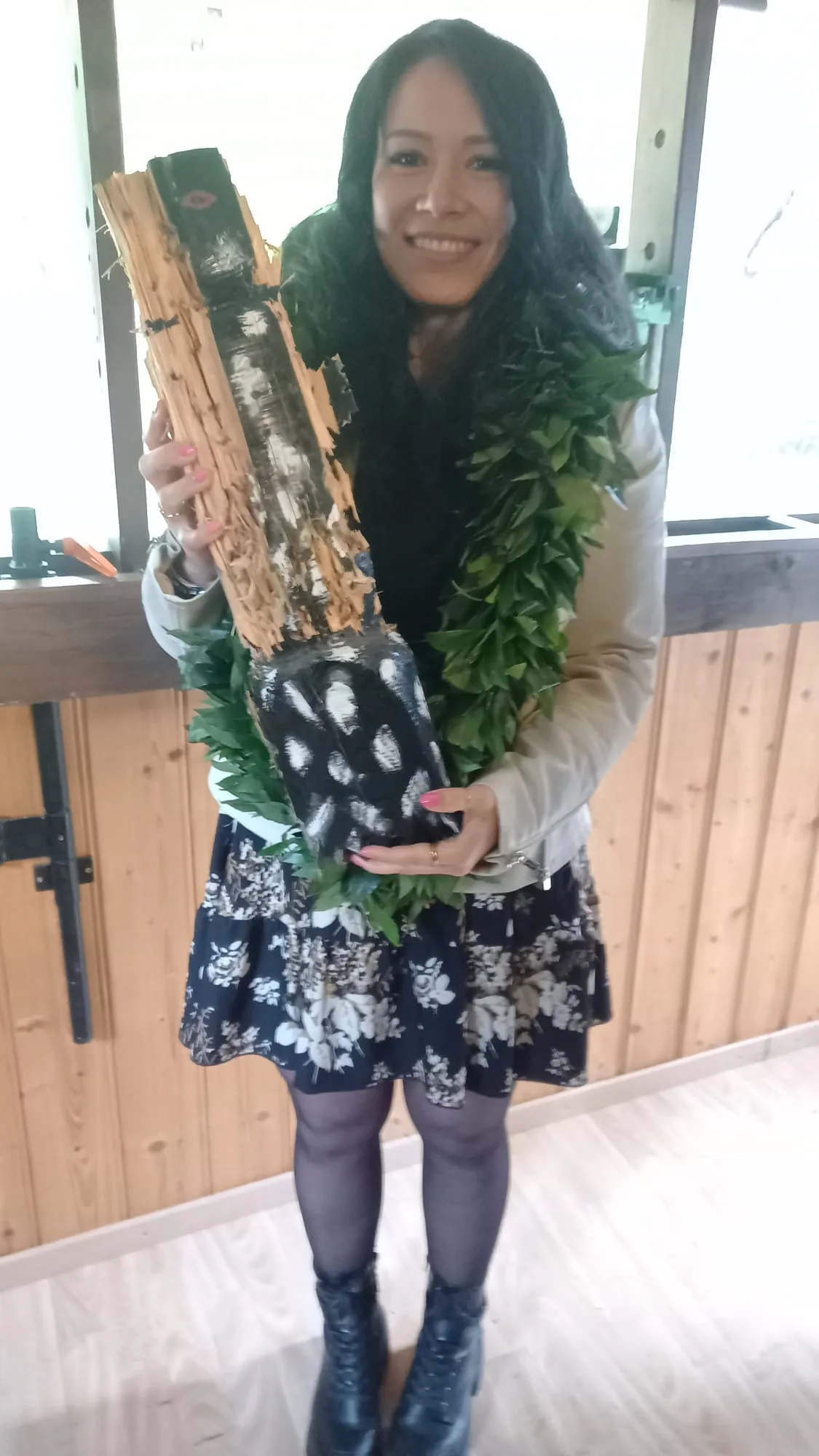 Eine junge Frau mit langen, dunklen Haaren lächelt in die Kamera und hält einen mit Blumen verzierten Behälter. Sie trägt einen beigen Mantel, ein geblümtes Kleid und schwarze Stiefel.