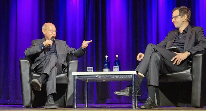 Lorenz Hemicker spricht in ein Mikrofon und zeigt auf Gregor Gysi während eines Gesprächs vor einem violetten Vorhang.
