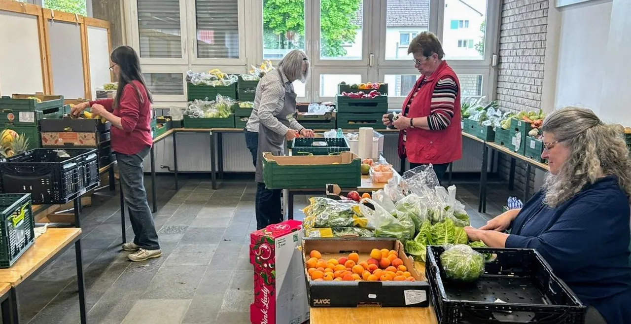 Vier Frauen arbeiten in einer großen Halle an Tischen mit Obst und Gemüse. Sie sortieren und verpacken Lebensmittel, die in Kisten und auf Paletten bereitstehen.