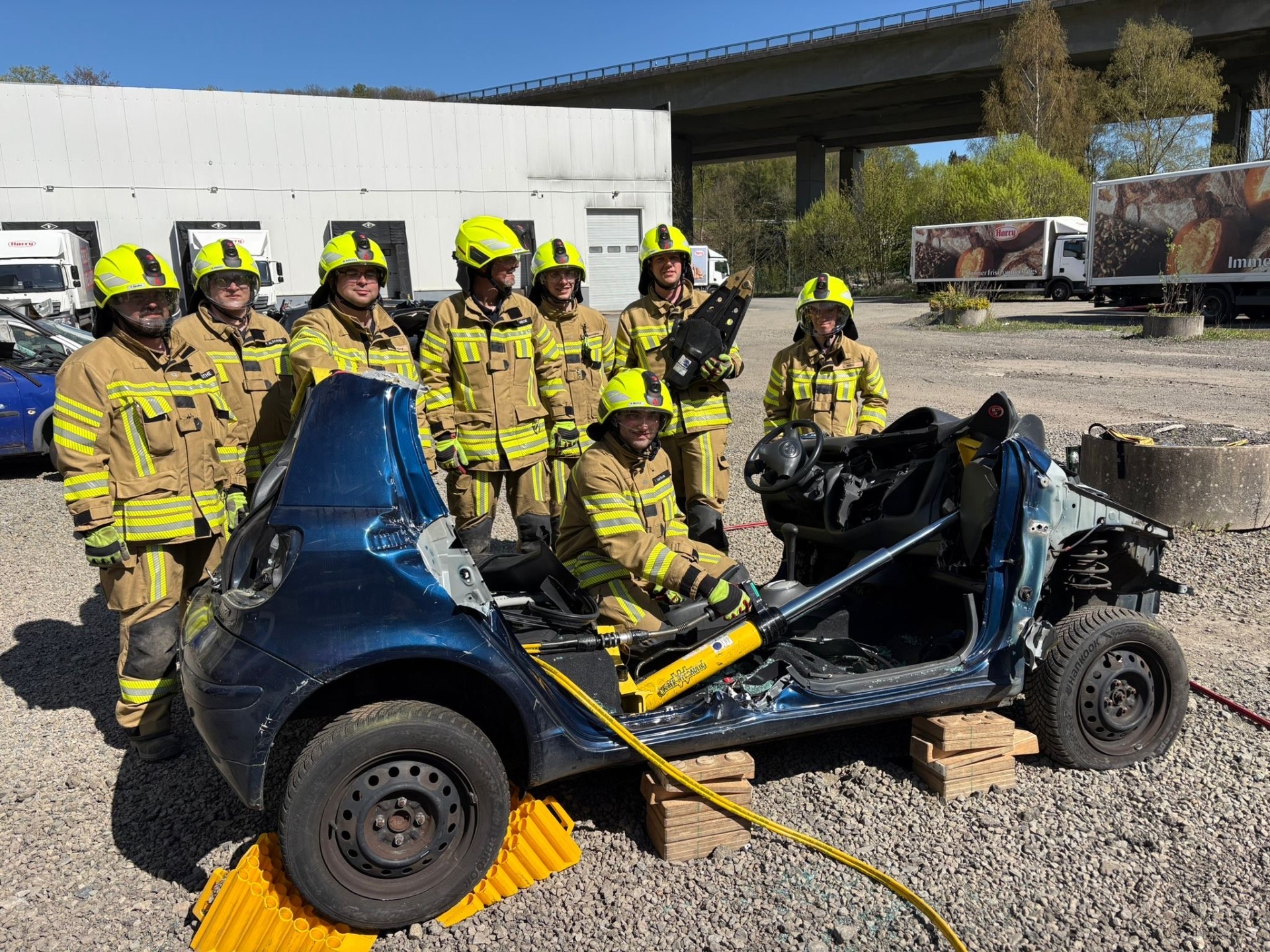 Acht Feuerwehrleute in voller Montur stehen um ein stark beschädigtes Auto herum, das auf einer Holzpalette steht. Im Hintergrund sind weitere Fahrzeuge und Industriegebäude zu sehen.