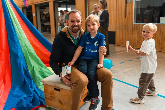 Ein Mann sitzt mit einem kleinen Jungen im blauen Trikot auf seinem Schoß. Im Hintergrund steht ein weiteres Kind und eine Gruppe von Menschen. Im Vordergrund ist ein Teil einer bunten Hüpfburg zu sehen.
