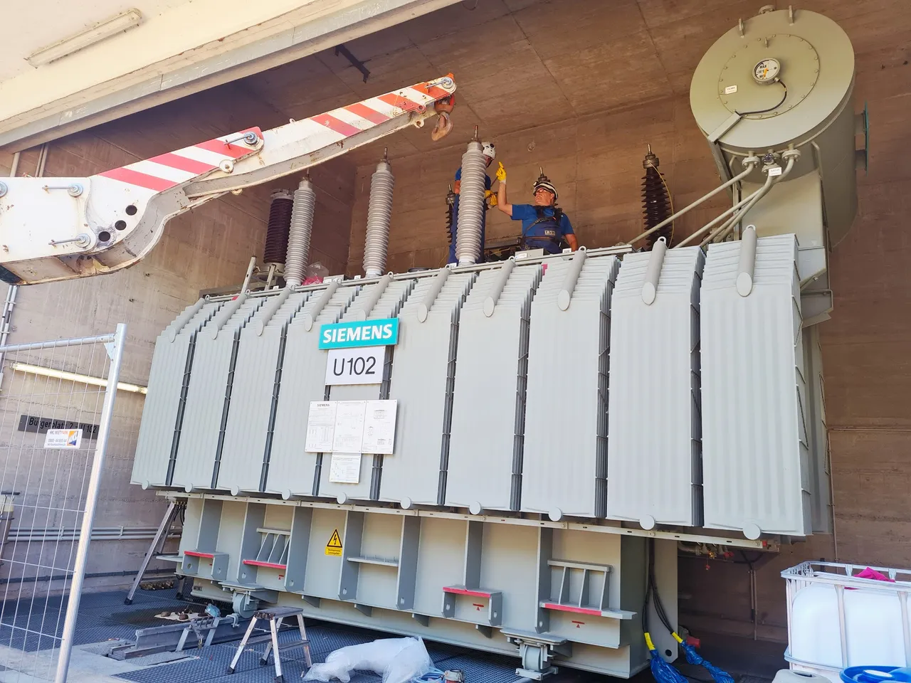 Zwei Arbeiter in Schutzkleidung stehen auf einem großen, grauen Siemens-Transformator (U102) unter einer Betonkonstruktion, während ein Kranarm darüber positioniert ist.