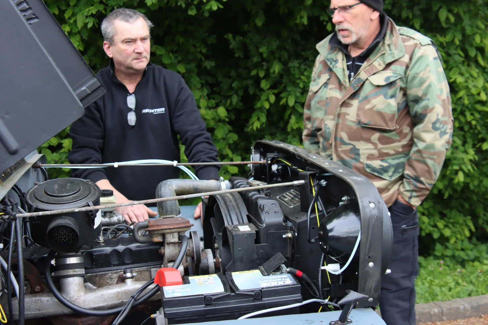 Zwei Männer betrachten den geöffneten Motorraum eines Oldtimers. Der Mann links trägt einen schwarzen Pullover, der rechte eine Jacke mit Tarnmuster.
