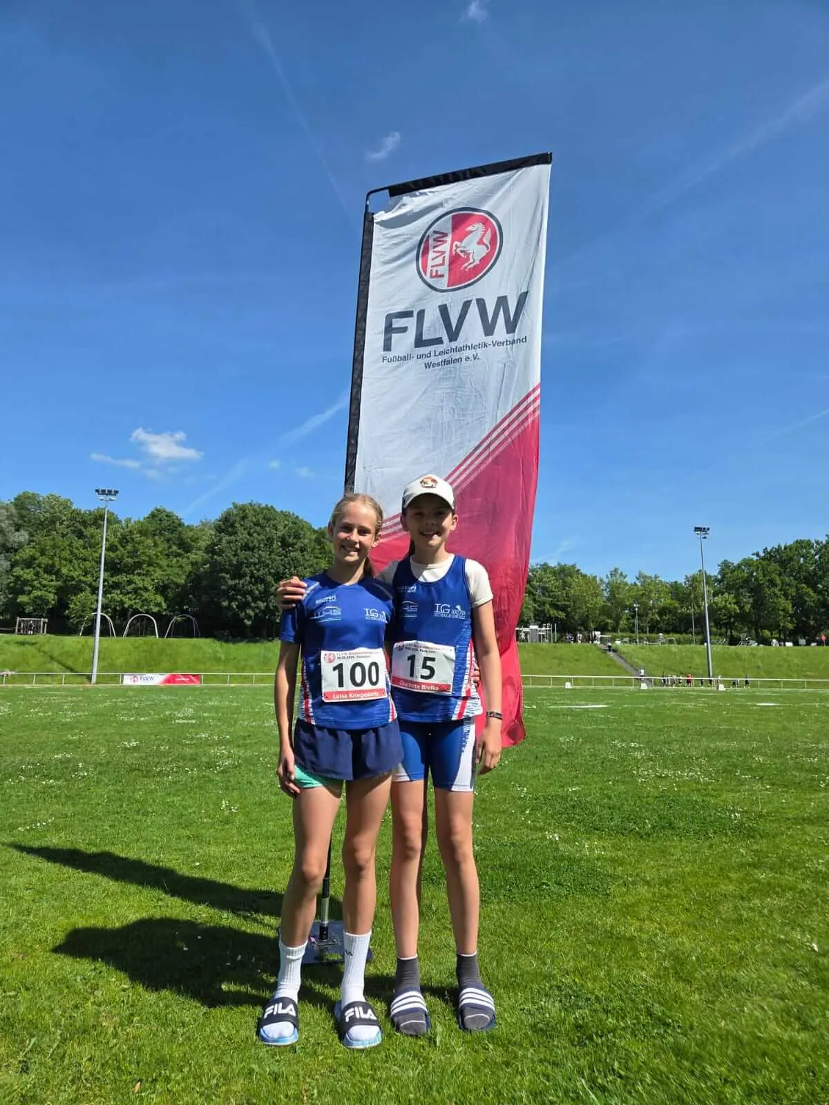 Zwei junge Läuferinnen mit Startnummern 100 und 15 stehen vor einer rot-weißen Flagge des FLVW (Fußball- und Leichtathletik-Verband Westfalen) auf einer grünen Wiese. Im Hintergrund eine Sportanlage und ein blauer Himmel.