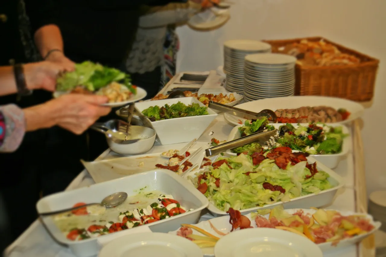 Ein Buffet mit verschiedenen Salaten, Brot und Tellern, eine Person hält einen Teller mit grünem Salat.