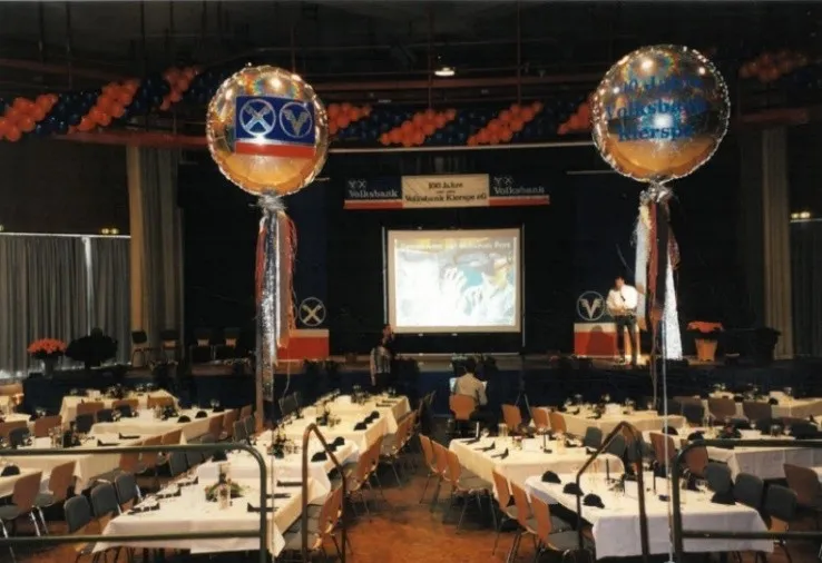 Festsaal mit festlich gedeckten Tischen, einer Bühne mit Redner und einer Präsentation auf einer Leinwand. Zwei große Ballons mit dem Logo der Volksbank Kierspe flankieren den Raum.