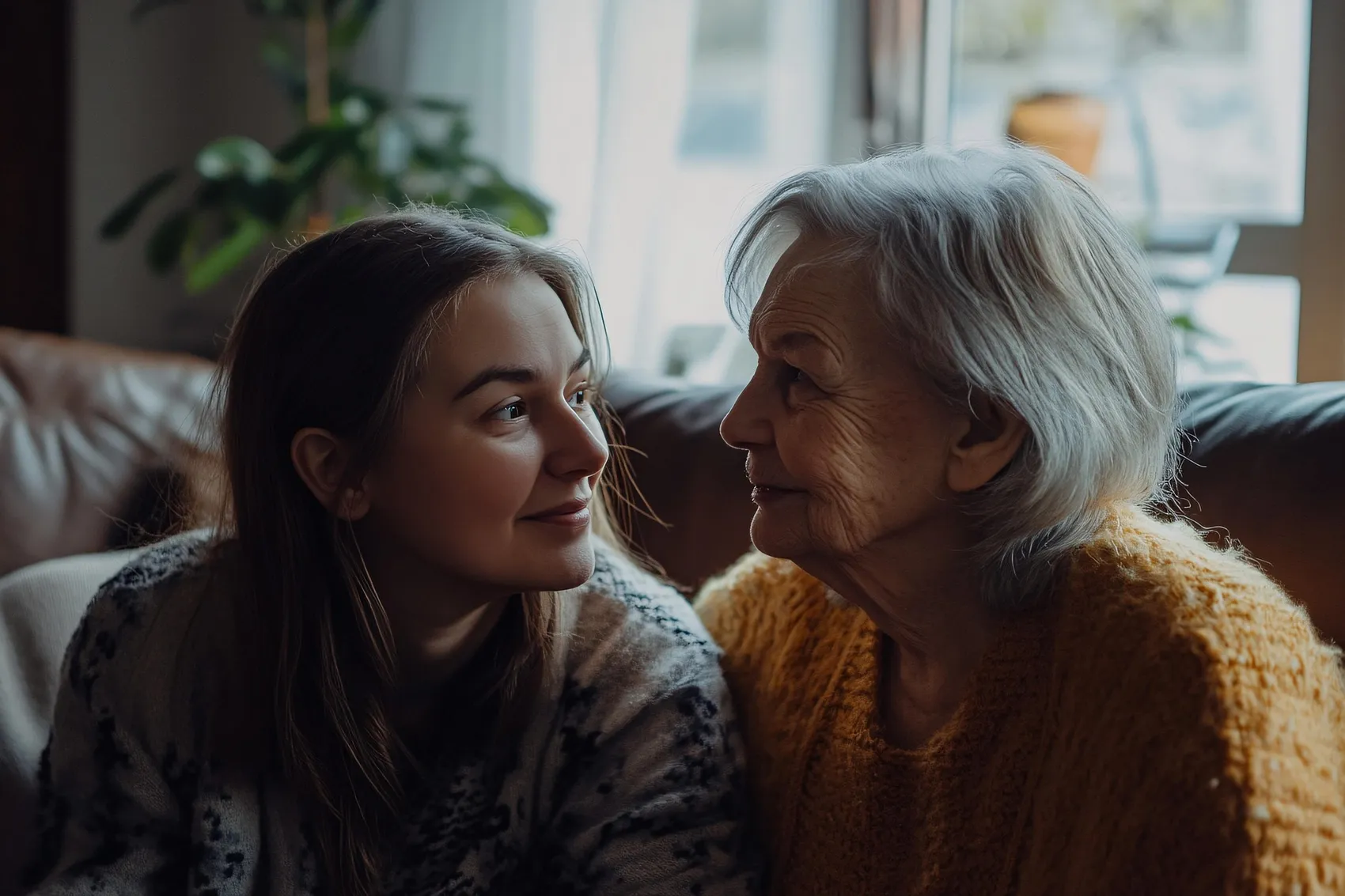 Junge Frau und ältere Frau blicken sich lächelnd zu, sitzen nebeneinander auf einem Sofa. Das Bild symbolisiert die Betreuung von Angehörigen und die damit verbundenen Herausforderungen.