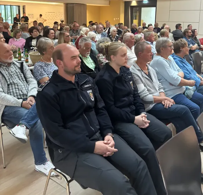 Ehrenamtliche in dunkelblauen Uniformen sitzen in den vorderen Reihen eines Saals, während im Hintergrund weitere Personen sitzen.