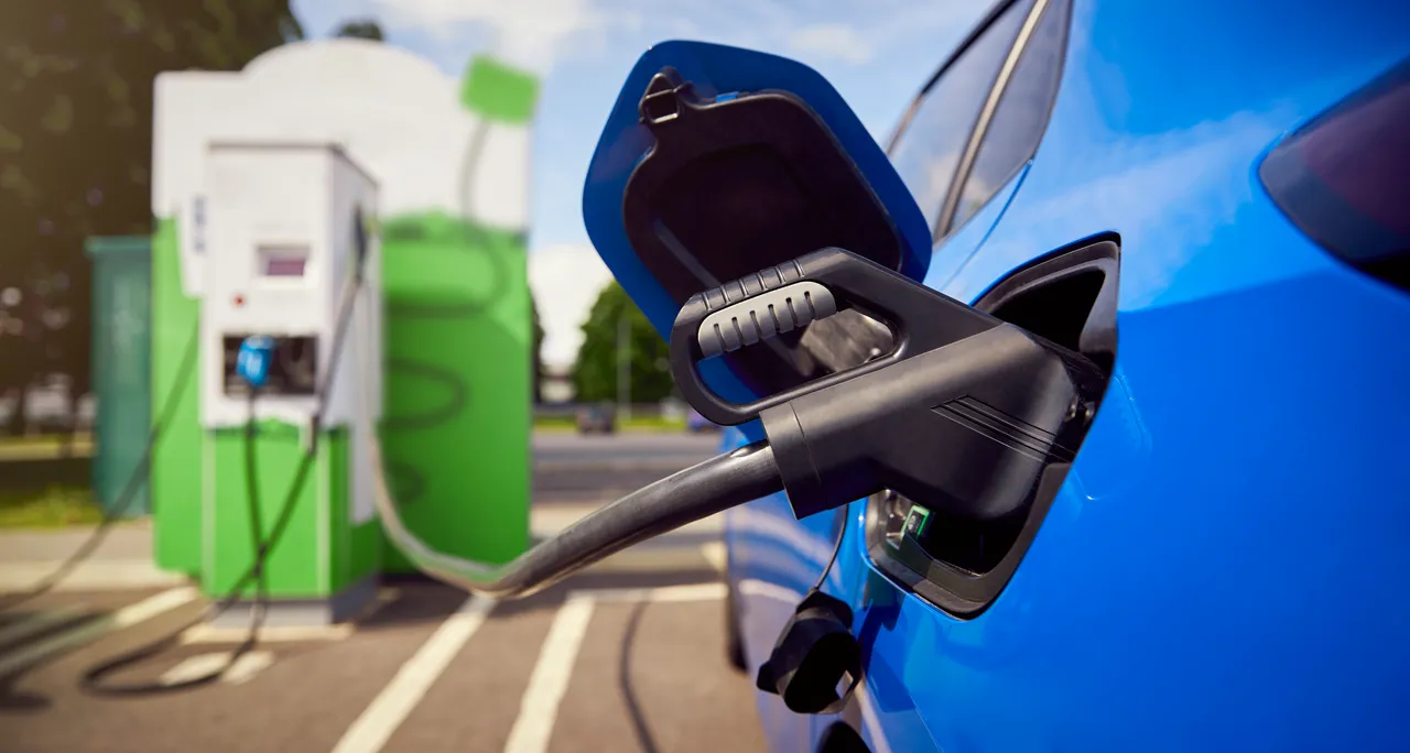 Ein schwarzes Ladekabel steckt im Ladeanschluss eines blauen Elektroautos vor einer grünen Ladestation.