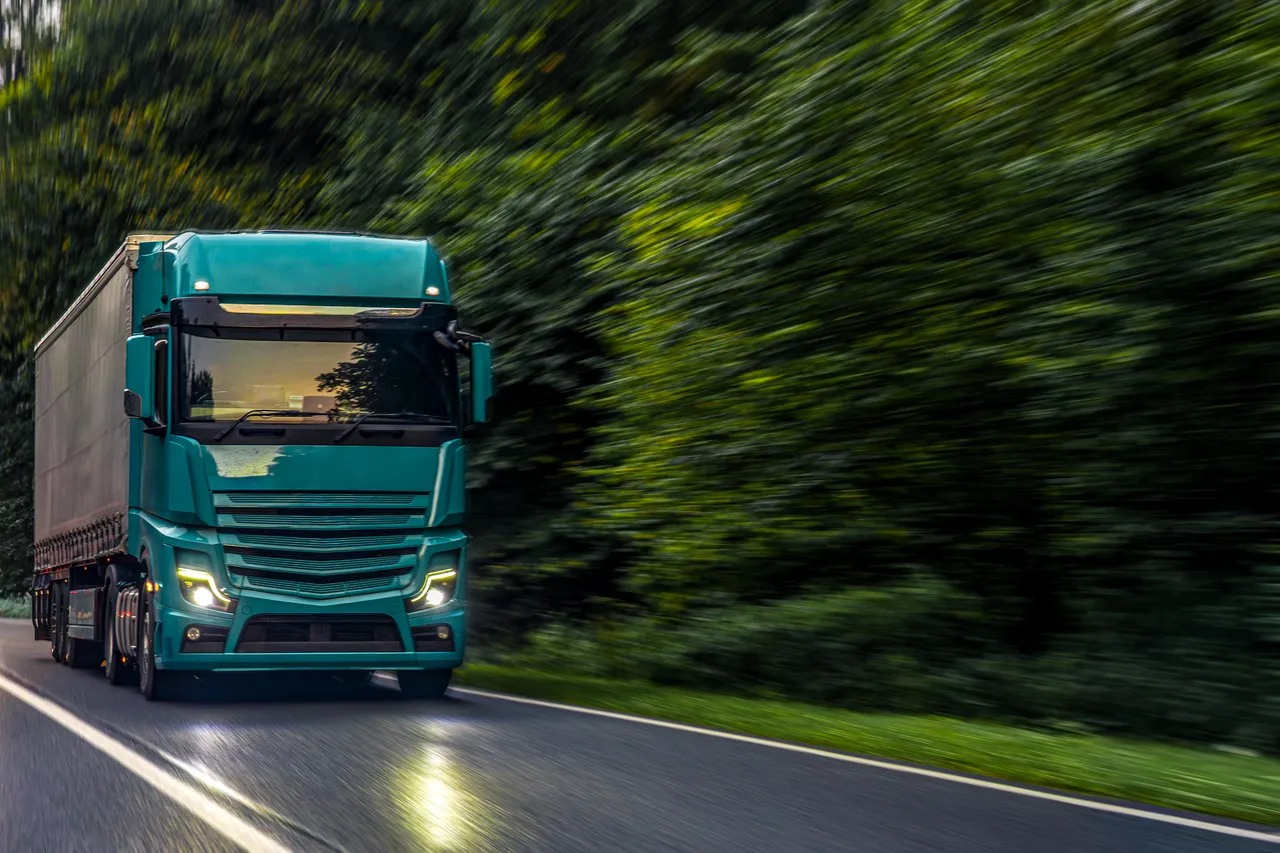 Grüner Sattelschlepper fährt auf einer Landstraße durch eine bewaldete Landschaft.