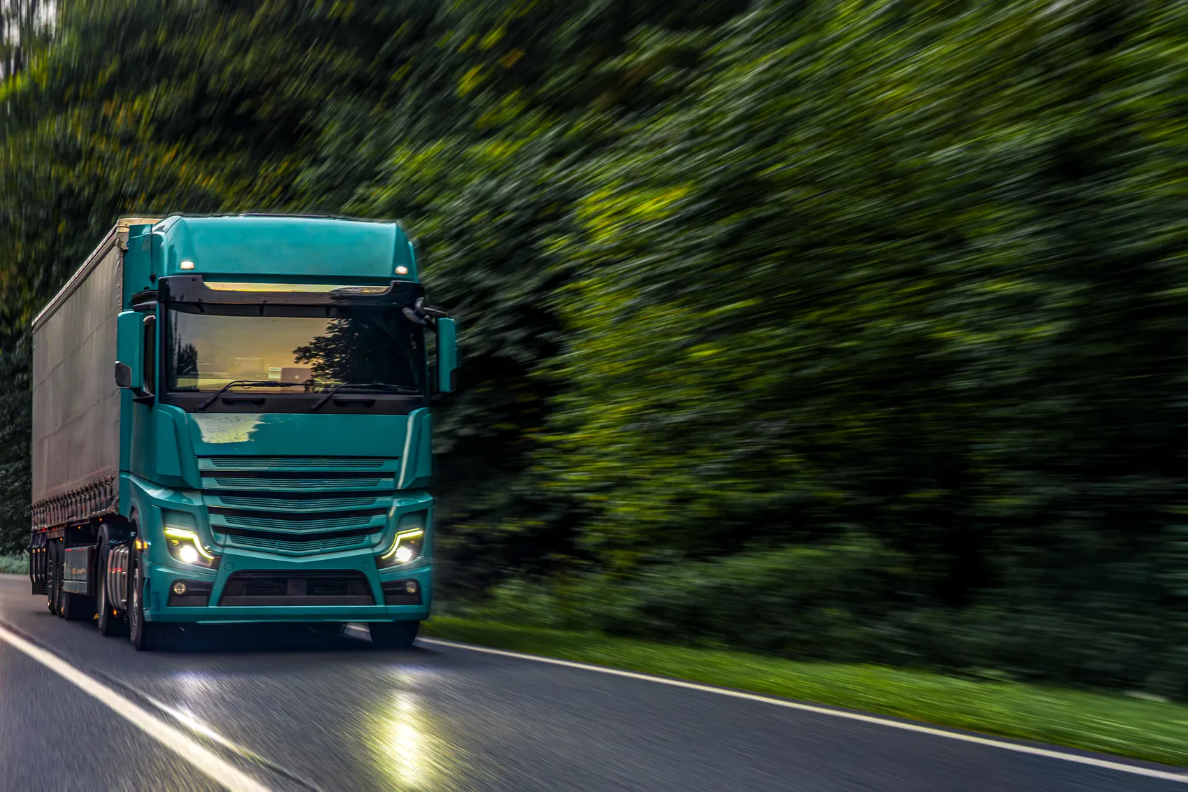 Grüner Sattelschlepper fährt auf einer Landstraße durch eine bewaldete Landschaft.