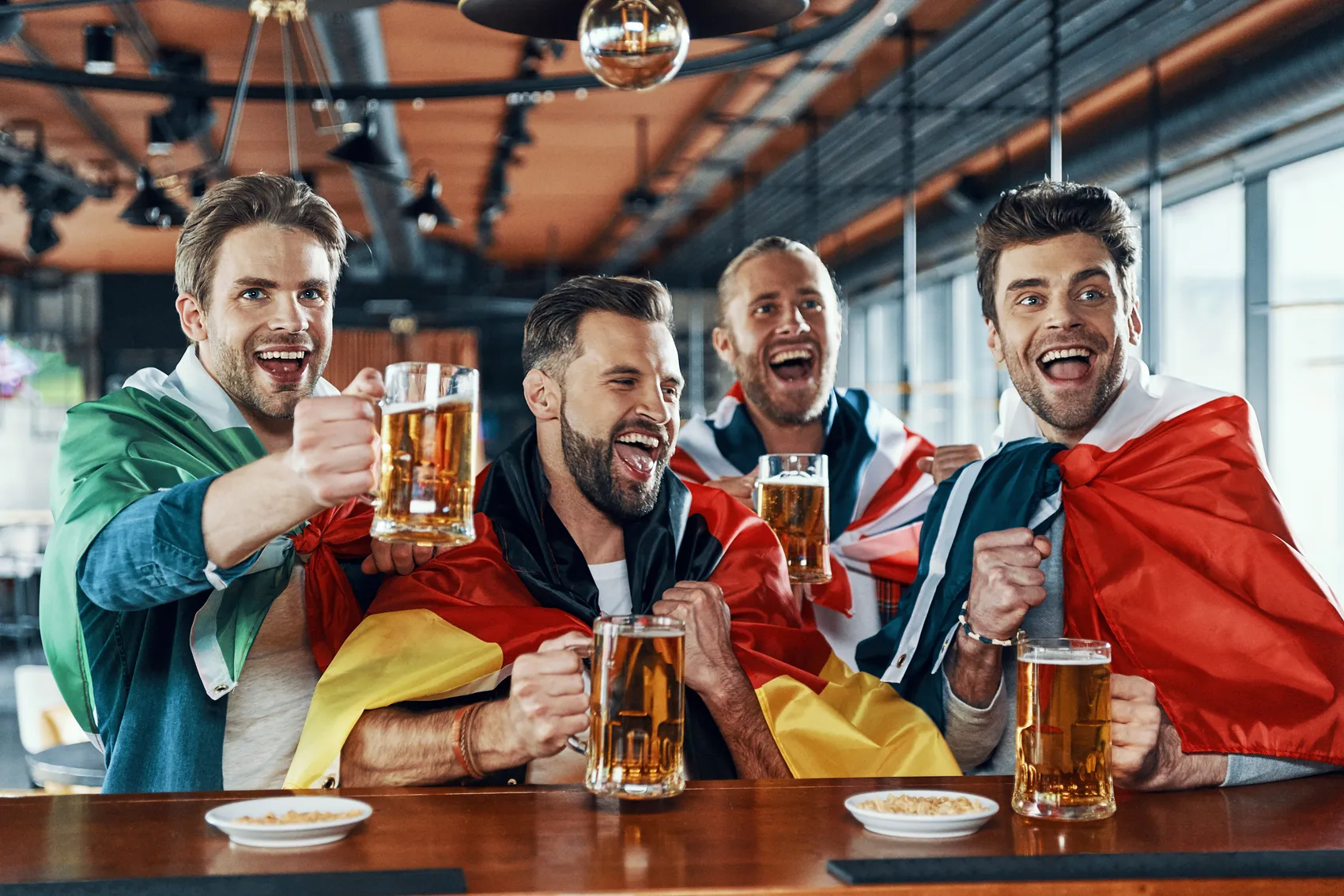 Vier Männer mit Bierkrügen stoßen in einer Bar an, umgeben von den Flaggen verschiedener Länder. Sie lächeln und jubeln, vermutlich während einer Fußball-WM-Übertragung.