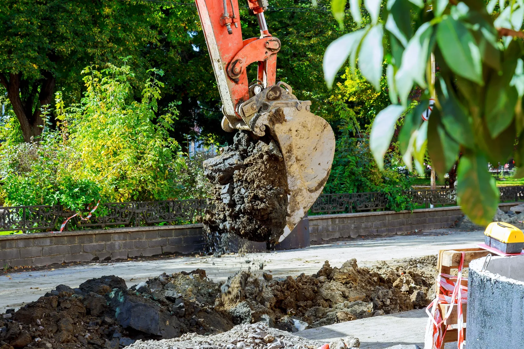 Bagger schaufelt Erde in eine Grube neben einer Baumreihe und einer Mauer. Im Vordergrund Baustellenabsperrung.