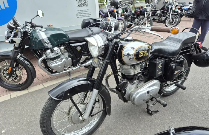 Zwei klassische Royal Enfield Motorräder stehen nebeneinander auf einem Pflasterweg, ein drittes Motorrad ist im Hintergrund zu sehen. Im Hintergrund ein blaues Werbebanner.