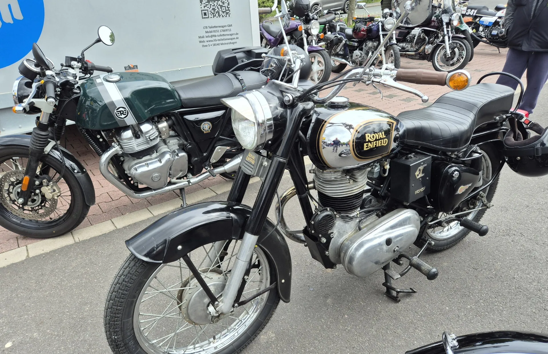 Zwei klassische Royal Enfield Motorräder stehen nebeneinander auf einem Pflasterweg, ein drittes Motorrad ist im Hintergrund zu sehen. Im Hintergrund ein blaues Werbebanner.