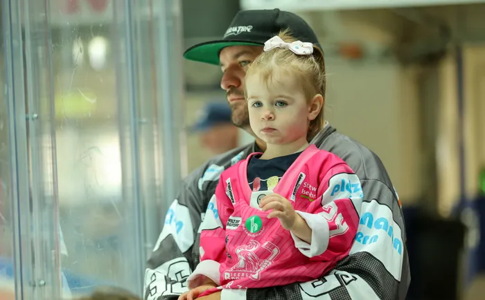 Ein Mann hält ein kleines Mädchen in einem rosa Iserlohn Roosters Trikot in einer Eissporthalle.