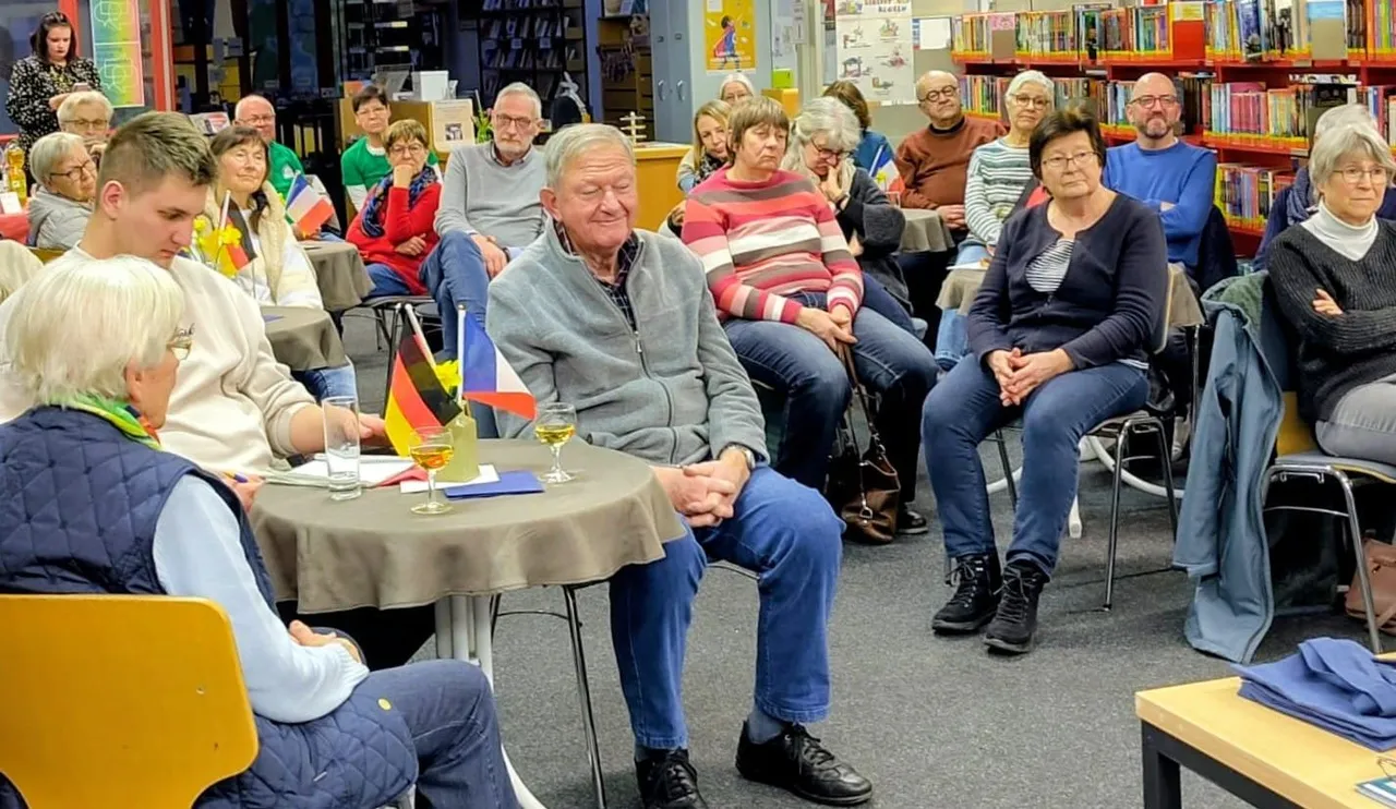 Zuschauer sitzen auf Stühlen und an Tischen in einer Bibliothek. Im Vordergrund ein Mann mit einem Glas Wein, im Hintergrund Regale mit Büchern.