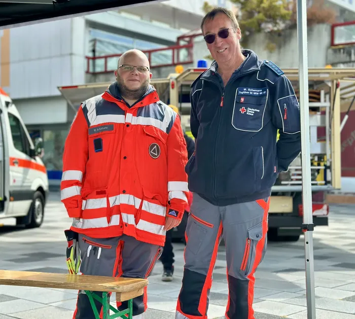 Zwei Männer in Uniformen des Deutschen Roten Kreuzes stehen im Freien; einer trägt eine orangefarbene Warnjacke, der andere eine dunkelblaue Jacke.