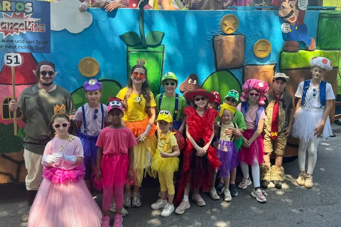Eine Gruppe von Kindern und Jugendlichen in bunten Kostümen steht vor einem farbenfrohen Hintergrund mit Mario-Motiven. Sie lächeln in die Kamera und halten eine Trophäe. Ein Mann und eine Frau stehen seitlich neben der Gruppe.