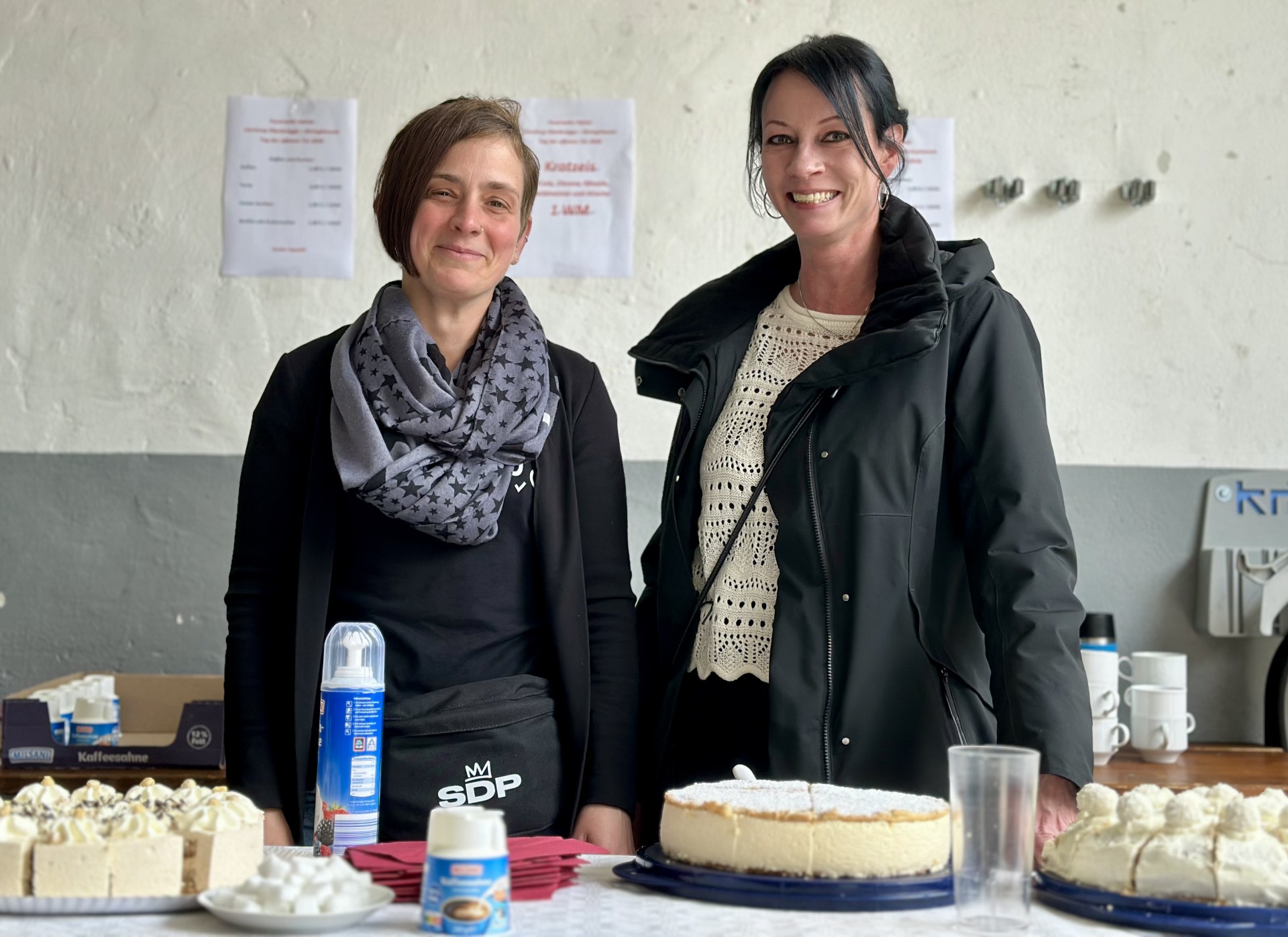 Zwei lächelnde Frauen stehen vor einem Tisch mit Kuchen und Gebäck. Im Hintergrund sind Plakate und eine Kaffeemaschine zu sehen.